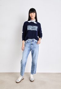 Maglione blu navy con testo bianco, jeans larghi azzurri chiari e sneaker argentati. Il modello indossa anche una sciarpa leggera annodata attorno al collo.