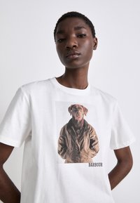 Bílý bavlněný tričko s potiskem obrázku psa v hnědém kabátu; text "BARBOUR" pod grafikou. Krátké rukávy a kulatý výstřih.