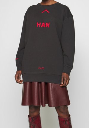 Osoba ubrana w za dużą czarną bluzę z czerwonymi literami "HAN", czerwoną plisowaną skórzaną spódnicę oraz czerwone botki w wężowy wzór stojąca na białym tle.