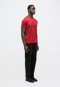 Rød T-shirt med korte ærmer, der har et sort logo-patch på venstre bryst, kombineret med sorte jeans og sorte sneakers.