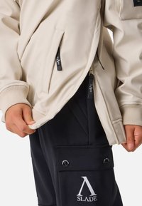 Giacca beige con polsini a coste, tasche con zip e dettagli con logo. Pantaloni scuri con tasca frontale e dettagli a bottone automatico. Texture liscia, design casual.