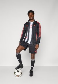Sort træningsjakke med røde striber, hvid skjorte, sorte shorts, sorte fodboldstøvler og sorte sokker med hvide striber, stående på en fodbold.