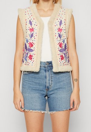Veste sans manches crème avec broderies florales violettes et roses et bordure en fourrure portée sur un haut blanc avec un short en jean bleu taille haute.
