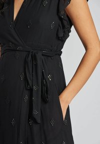 Robe noire enroulée avec des manches volantes, une taille à nouer, et des accents brodés complexes avec des embellissements brillants. Tissu texturé.