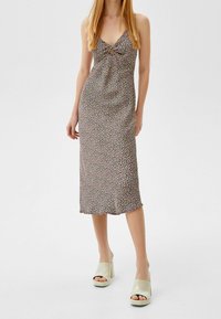 Kobieta w kwiecistej, bezrękawnikowej sukience midi z detalem w postaci węzła z przodu oraz beżowych sandałach na obcasie z odkrytymi palcami na białym tle.