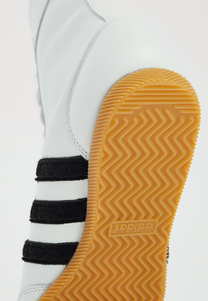 Weißer Sportschuh mit schwarzen Wildlederstreifen, ausgestattet mit einer strukturierten, orangefarbigen Gummisohle und einem glatten Lederobermaterial.