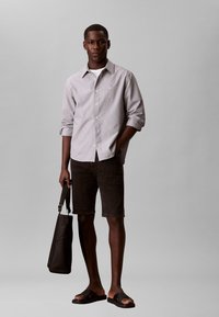 Άνδρας με ανοιχτό γκρι πουκάμισο με κουμπιά, μαύρο σορτς, μαύρα σανδάλια, κρατώντας μια μαύρη τσάντα, στέκεται μπροστά σε απλό γκρι φόντο.