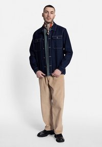 Casaco de ganga azul escuro com bolsos no peito, usado sobre uma camisa verde, combinado com calças de bombazina bege e sapatos pretos.