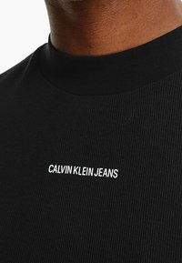 Svart ribbad t-shirt med hög hals och vitt broderat "CALVIN KLEIN JEANS"-logotyp på bröstet.