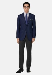 Námornícky blejzer s dvoma gombíkmi, nosený cez svetlomodrú košeľu a tmavú kravatu. Sivé upravené nohavice a čierne topánky dopĺňajú celý outfit.