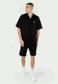 Camisa preta de botão de mangas curtas com colarinho, combinada com calções pretos. Detalhe do logótipo em branco. Modelo a usar ténis brancos e meias.