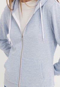 Sudadera con cremallera de color azul claro, hecha de tela suave, con una capucha forrada en blanco, bolsillos frontales y cordones, con una textura lisa.