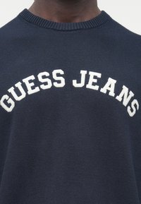 Tumši zils džemperis ar ribotu apkakli, priekšpusē krūtīs baltā krāsā izšūts uzraksts "GUESS JEANS". Materiālam ir gluda tekstūra.