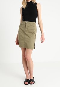 Jupe en coton kaki au design ajusté avec une fente sur le côté, associée à un débardeur noir côtelé et des sandales plates noires.