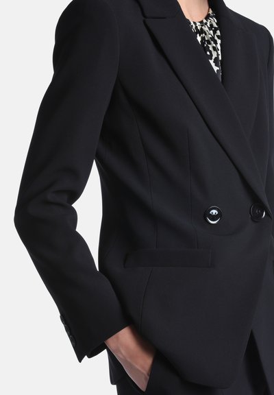 Blazer noir cintré à double boutonnage avec revers à pointes porté sur un top noir et blanc à motifs, main placée dans la poche latérale.