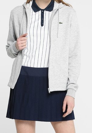 Vrouw draagt een grijze hoodie met rits over een wit poloshirt met marineblauwe strepen en een marineblauwe geplooide rok, staand tegen een effen achtergrond.