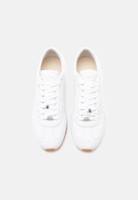 Paire de baskets basses blanches à lacets avec semelles marron clair, vues de dessus sur un fond gris clair uni.