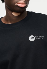 Μαύρο βαμβακερό φούτερ με στρογγυλή λαιμόκοψη, με λευκό κυκλικό λογότυπο και το κείμενο "New Balance Athletics Inc." στην περιοχή του στήθους.