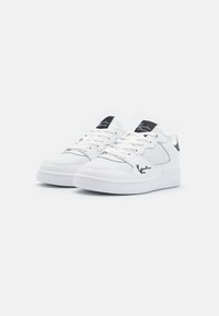 Hvide sneakers med en tekstureret overflade, perforerede sektioner og sorte detaljer på hælen og tungen. Rundt tå og flad sål.