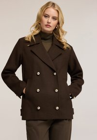 Brązowy dwurzędowy płaszcz z wełny, z połyskującymi srebrnymi guzikami, szerokimi klapami i kieszeniami po bokach; noszony na zielonym golfie.