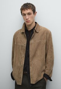 Tan suede button-up jacket with a collar, featuring a smooth texture and snap closures. Layered over a dark checkered shirt.