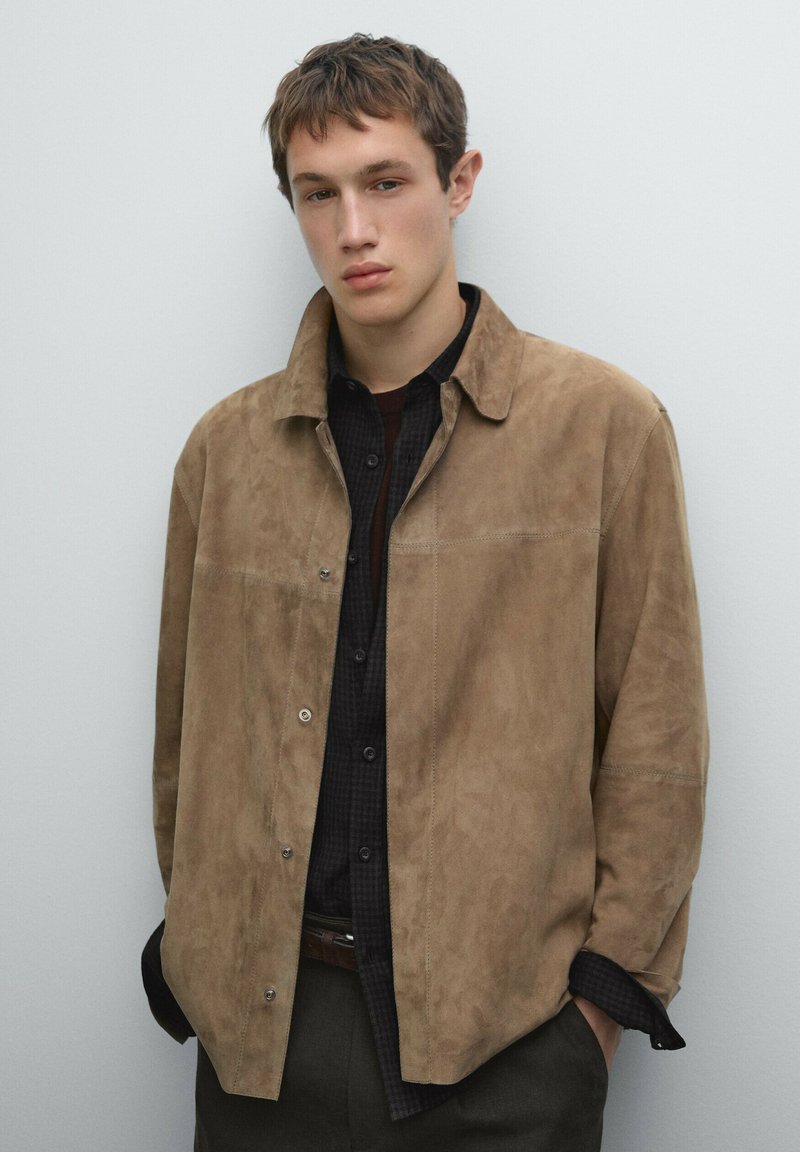 Tan suede button-up jacket with a collar, featuring a smooth texture and snap closures. Layered over a dark checkered shirt.