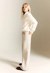 Kerma ribbed-neule, vaaleanbeige leveälahkeiset housut sivusaumalla ja tummanruskeat kengät. Malli seisoo kädet taskuissa ja katsoo suoraan eteenpäin.
