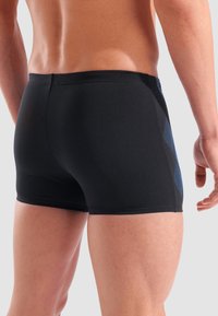 Schwarze Schwimmshorts mit glatter Textur, die ein blaues diagonales Muster auf einer Seite aufweisen, in mittellanger Länge bis zur Oberschenkelmitte und mit tailliertem Schnitt.