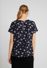 Navyfarbene Bluse mit kurzen Ärmeln, ausgestattet mit einem Reißverschluss auf der Rückseite, kleinen rosa und weißen Blumenmustern sowie einer leichten, glatten Textur.