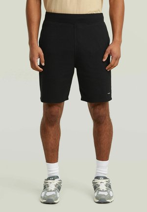 Uomo che indossa pantaloncini neri testurizzati, calzini bianchi e scarpe da ginnastica grigie, in piedi su uno sfondo neutro.