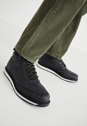 Černé kožené turistické boty s bílými podrážkami nošené s olivově zelenými manšestrovými kalhotami na bílém pozadí.