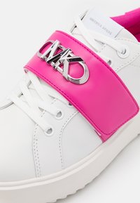 Sneaker in pelle bianca con cinturino lucido rosa e dettaglio del logo metallico. Presenta lacci bianchi e una suola testurizzata per una migliore aderenza.