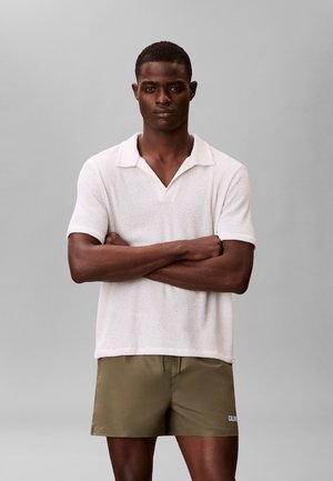 Homme aux bras croisés portant un polo à manches courtes blanc texturé et un short vert olive, sur fond gris uni.