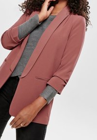Rose-colored blazer made of smooth fabric, featuring a lapel collar, rolled sleeves, and a single button closure. Worn over a gray striped top.