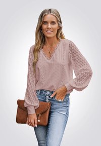 Femme avec de longs cheveux blonds portant un blouse en dentelle rose, un jean taille haute bleu clair, des colliers en or, et tenant une pochette marron.