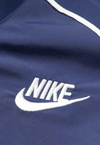 Tengerészkék szövet fehér Nike logóval. A hímzett szöveg vastag betűket és ikonikus pipa formátumot tartalmaz, sima textúrájú, matt felülettel.