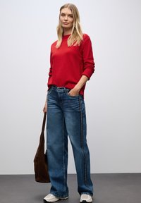 Maglione rosso con maniche lunghe, abbinato a jeans blu a gamba larga con strisce bianche ai lati. Il modello tiene una borsa marrone ed indossa scarpe bianche.