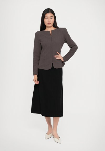 Woman with long dark hair wearing a textured gray zip-up jacket, black midi skirt, and white pointed shoes against a plain background.