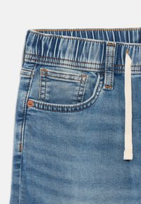 Pantaloni jogger din denim albastru deschis cu talie elastică, șnur și buzunar frontal. Prezintă cusături vizibile și o textură ușor estompată.