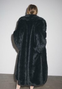 Crna lažna krzna jakna s širokim ovratnikom, velikim rukavima i teksturiranom, pahuljastom površinom, koja seže do sredine lista na figuri modela.