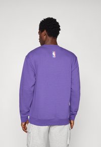 Sweatshirt roxo com mangas longas, decote redondo e punhos canelados. Apresenta um pequeno logótipo da NBA na parte de trás. O tecido parece suave e confortável.