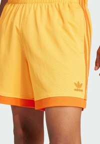 Yellow mesh sports shorts with orange accents and an elastic waistband. Features three orange stripes on the side and a small logo.
