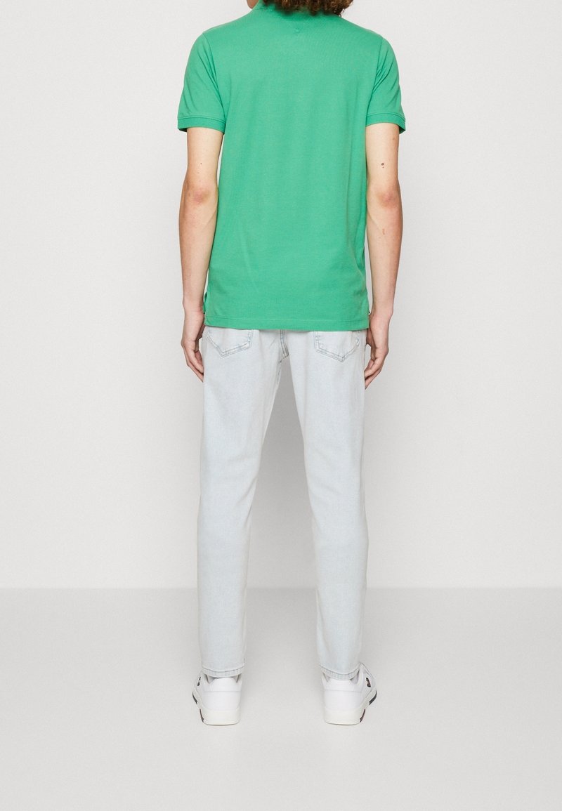 T-shirt vert en coton à manches courtes, porté avec un jean bleu clair et des baskets blanches. Le dos présente un design simple et un col rond.