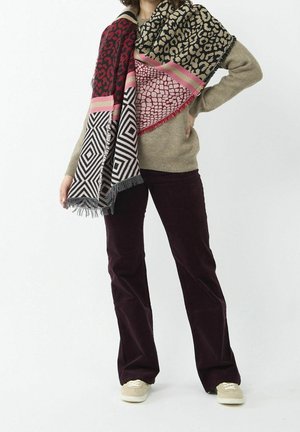 Personne portant un pull beige, un pantalon marron foncé, des baskets beiges et une écharpe oversize multicolore avec des motifs géométriques et animaliers.