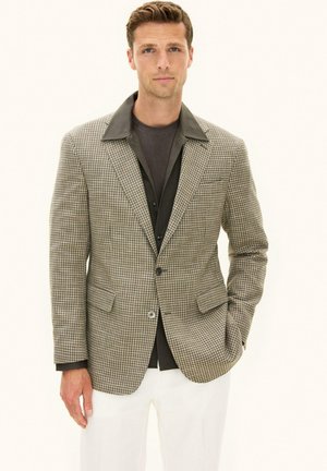 Man draagt een bruin houndstooth colbert over een donker shirt en witte broek, staat met een hand in de broekzak tegen een effen achtergrond.