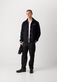 Navy zilu bumbvedēja jaka ar baltu apdari un logo, uzvilkta virs gaišas krekla, kombinēta ar melnām džinsām un melnu apavu, tur rokās kameru.