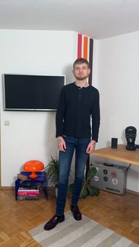 Svart långärmad henley-tröja och mörka jeans; tatueringar syns på händerna; glansiga bruna skor; inomhusmiljö med en orange lampa och en TV.