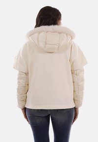 Giacca color crema con cappuccio foderato in pelliccia morbida, corpo in tessuto soffice e maniche trapuntate e lucide. Vista posteriore che mostra una vestibilità comoda e cuciture.