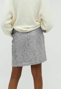 Mini-jupe argentée avec un motif vertical texturé, confectionnée dans un matériau scintillant. Associée à un pull crème oversize.