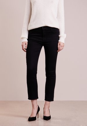 Person iført sorte slim-fit bukser, hvid ribstrikket sweater og sorte højhælede sko, stående på en lys gulv mod en neutral baggrund.